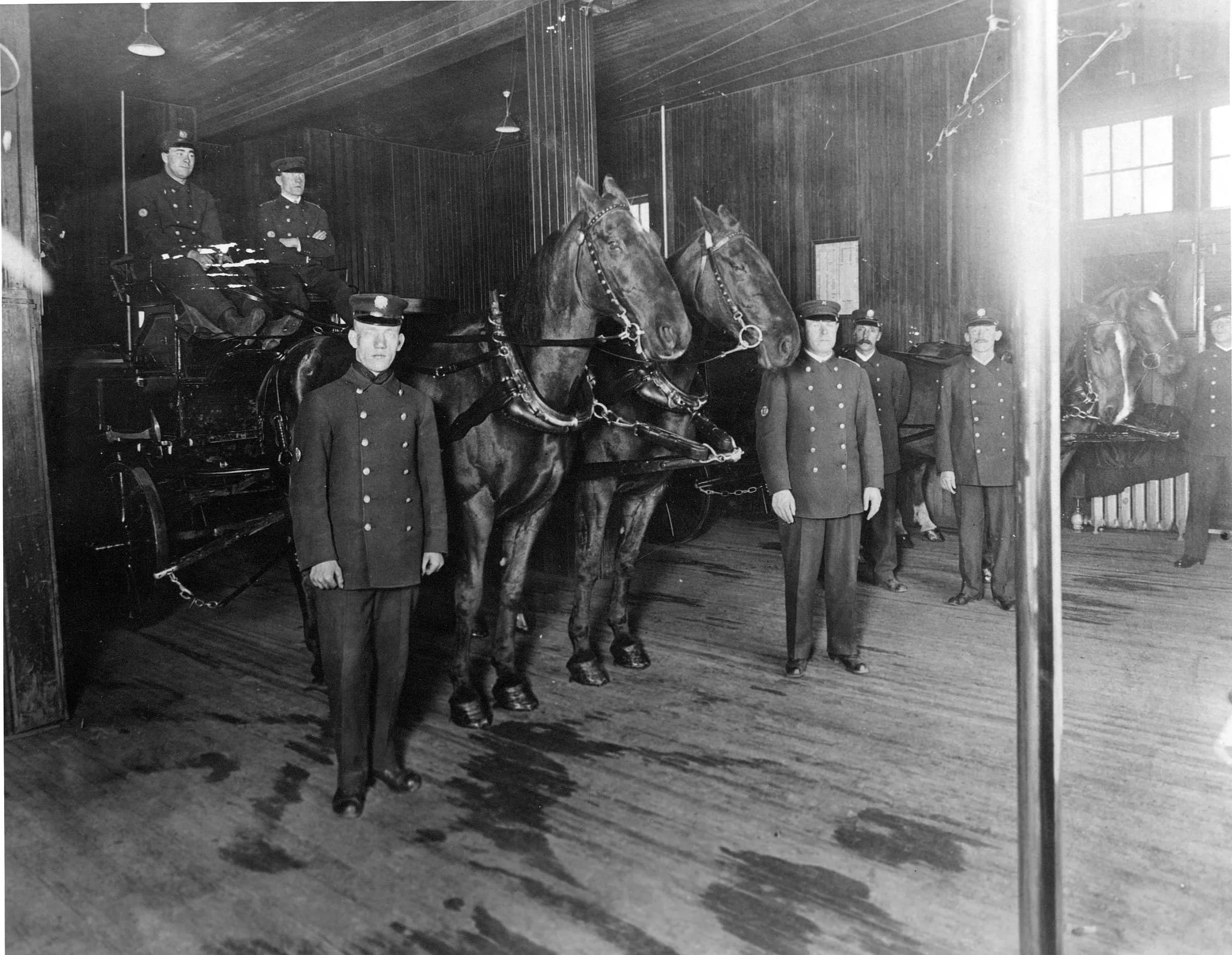 Uniformed firefighters stand with harnessed horses inside a fire hall.